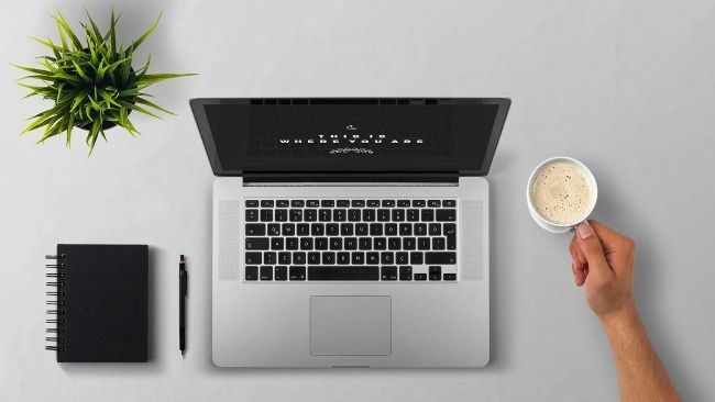 Laptop on desk with a hand holding a cup of coffee beside it