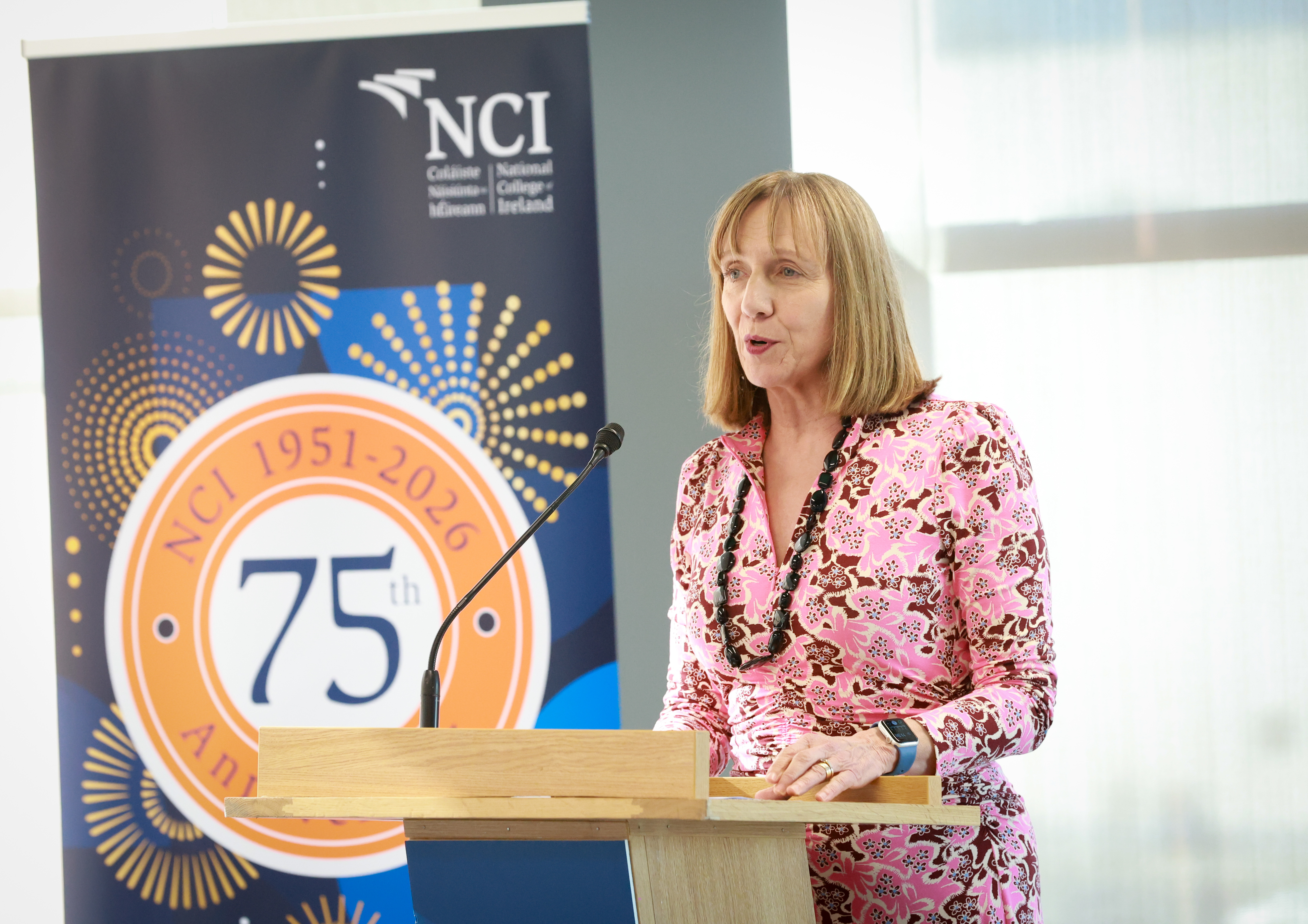 Professor Gina Quin speaking at a lectern in front of a sign for NCI's 75th anniversary